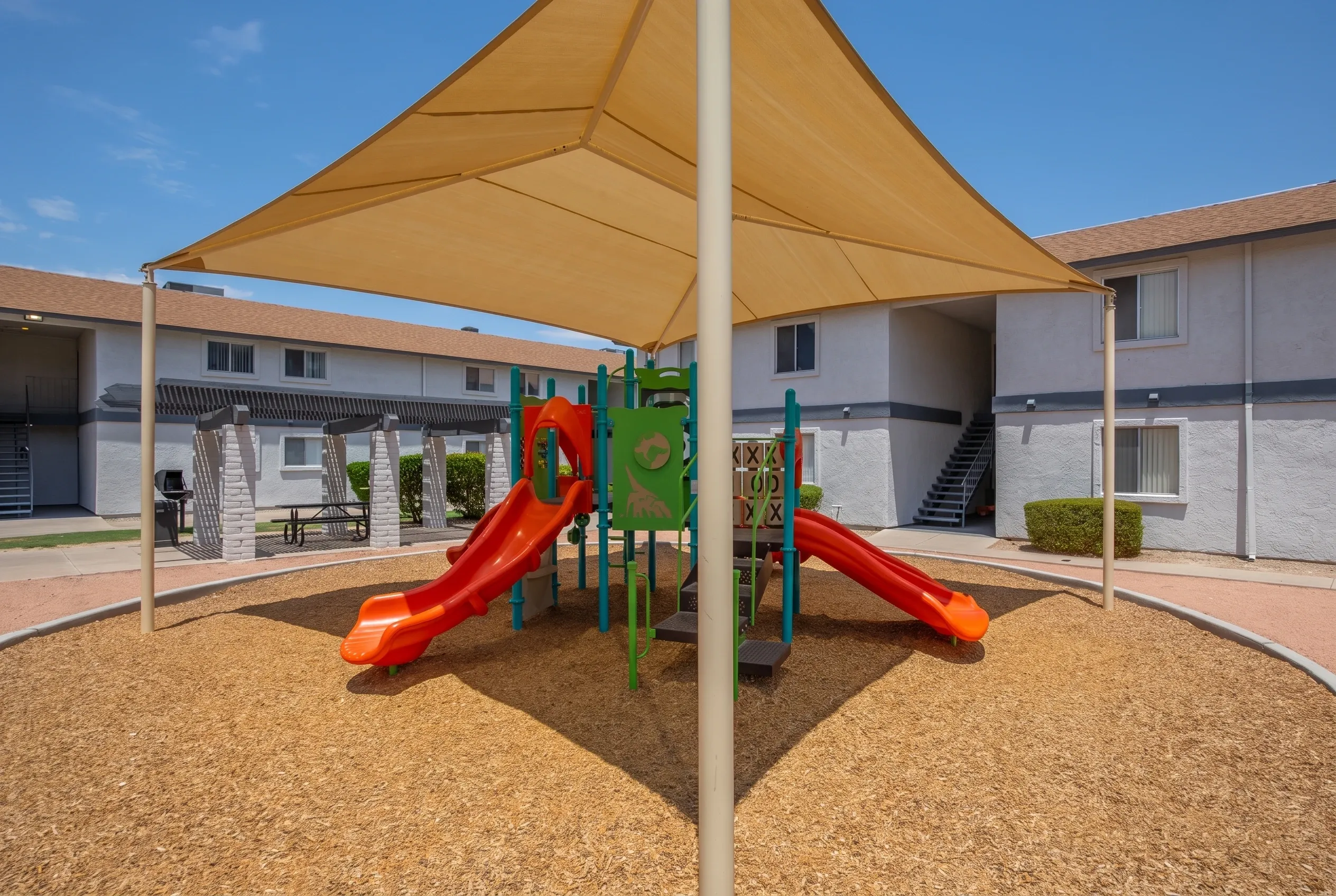 Playground area, red slides, green climbing structure, tan shade sail, wood-style mulch ground cover