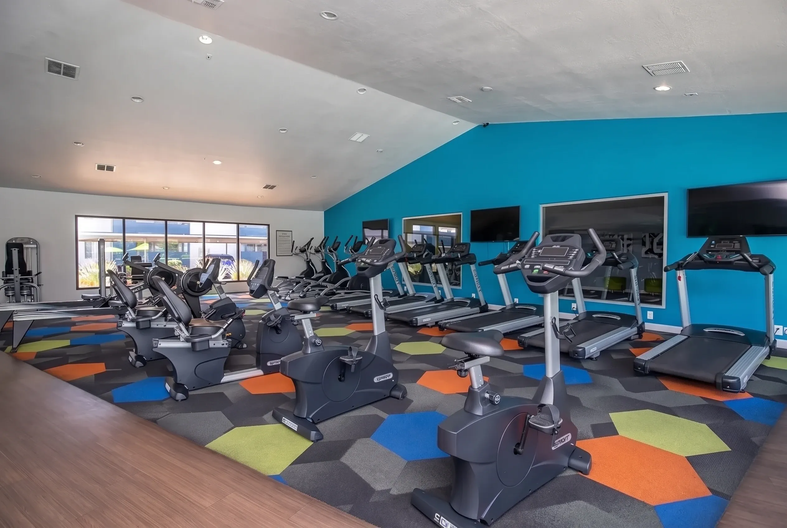Fitness center, stationary bikes and treadmills, turquoise accent wall, wood-style plank flooring, colorful floor tiles