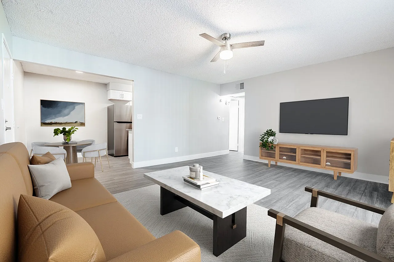 Living room with wood-style plank flooring, ceiling fan, wall-mounted TV, tan upholstered seating, open kitchen with stainless appliances visible