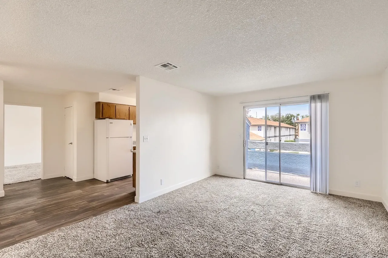 Living room, wood-style plank flooring, gray carpeted area, sliding glass patio doors, kitchen with cabinetry