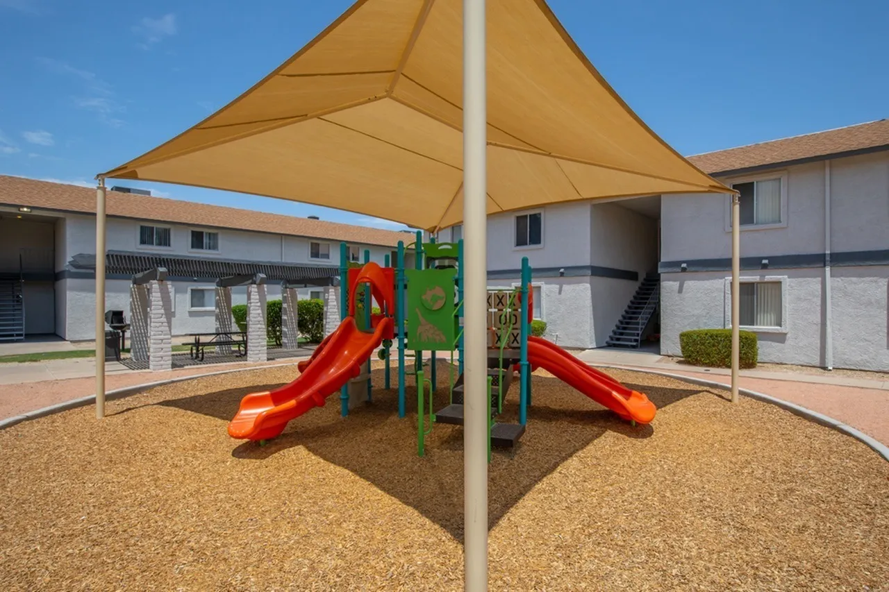 Playground area, red slides, green climbing structure, tan shade sail, wood-style mulch ground cover