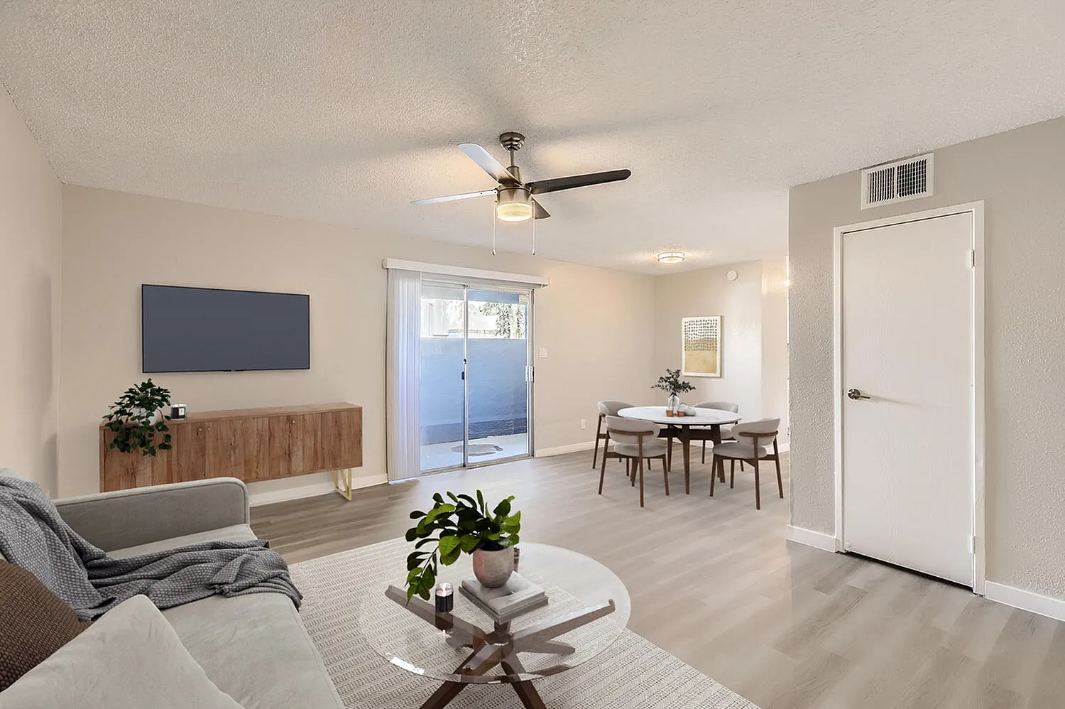 Living room with wood-style plank flooring, ceiling fan, sofa, round coffee table with plant, flat-screen TV mounted on wooden media console, dining area with table and chairs, glass sliding door to patio