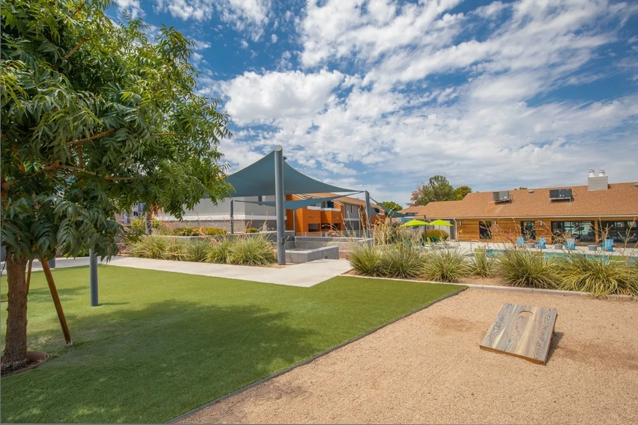 Community grounds with manicured lawn, shade trees, modern building structure, cornhole game, brick buildings