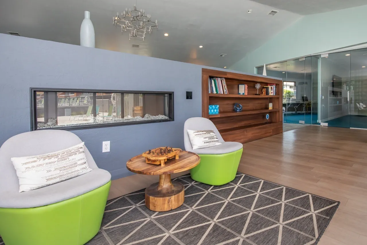 Community lounge, green barrel chairs, wood-style plank flooring, geometric area rug, built-in fireplace