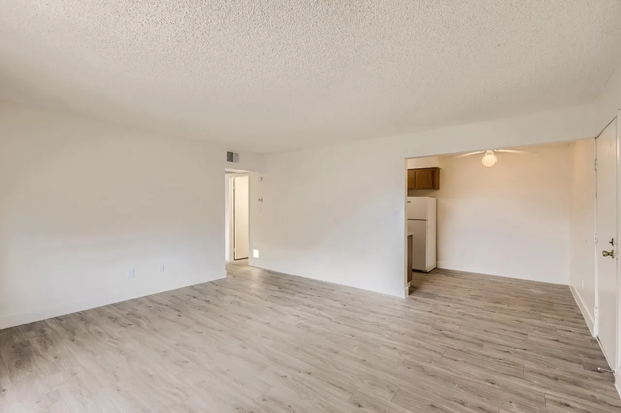 Living room, wood-style plank flooring, white walls, entry door, kitchen area