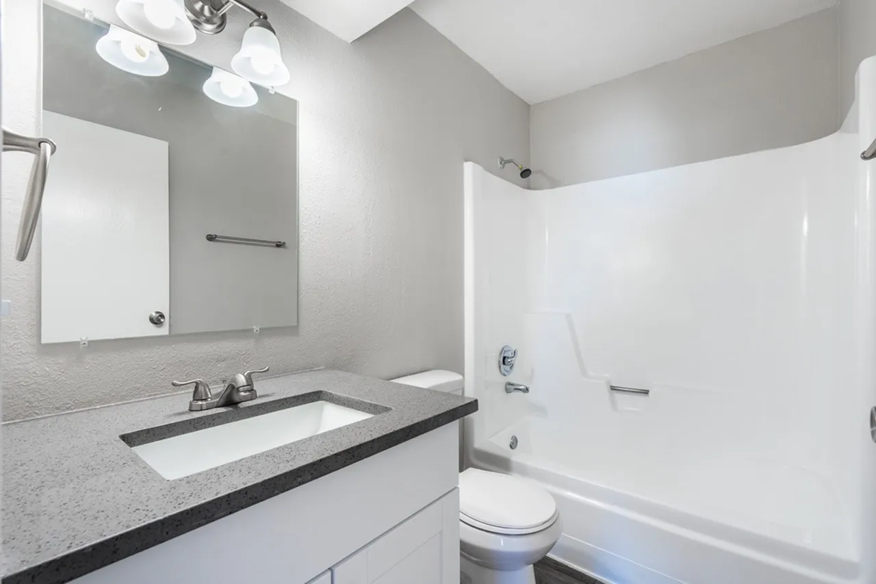 Bathroom with gray quartz countertops, white cabinetry, mirror and vanity lighting, white subway tile shower surround, toilet
