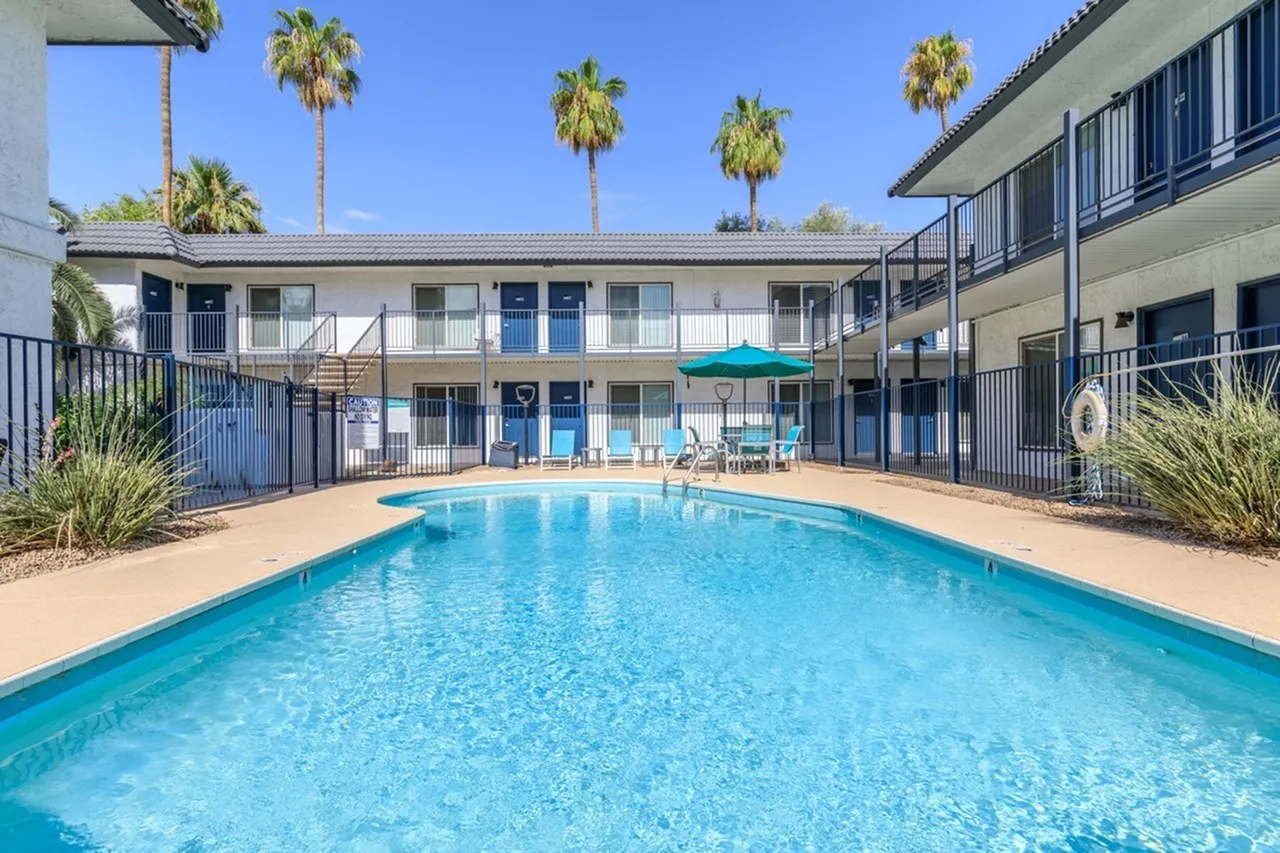 Resort-style swimming pool, tan concrete decking, lounge chairs, turquoise umbrella, multi-story residential building, palm trees