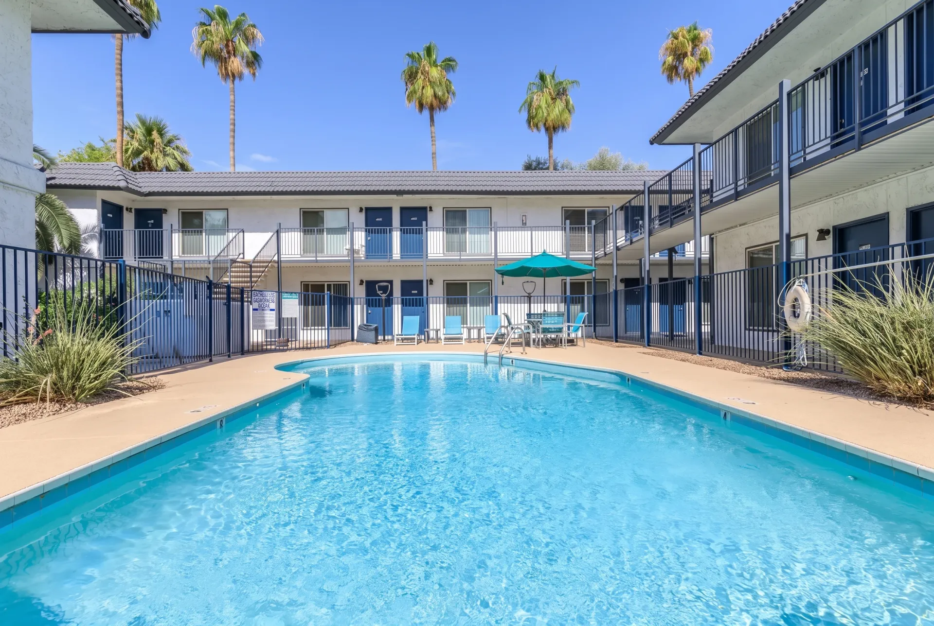 Resort-style swimming pool, tan concrete decking, lounge chairs, turquoise umbrella, multi-story residential building, palm trees