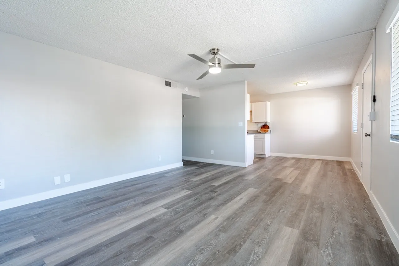 Living room, wood-style plank flooring, ceiling fan, white walls, entry door