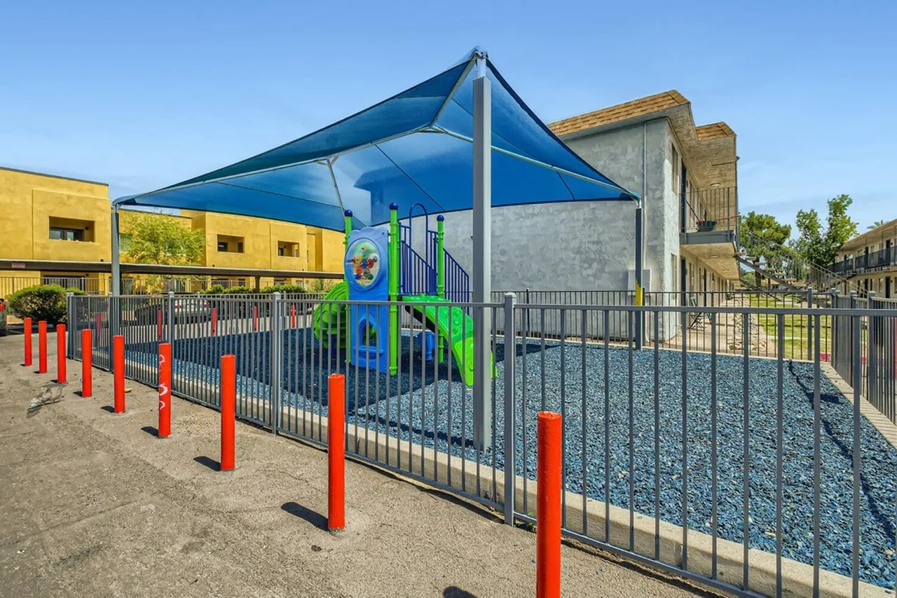 Playground with blue shade canopy, green play structure, red safety bollards, black fencing, gravel surface