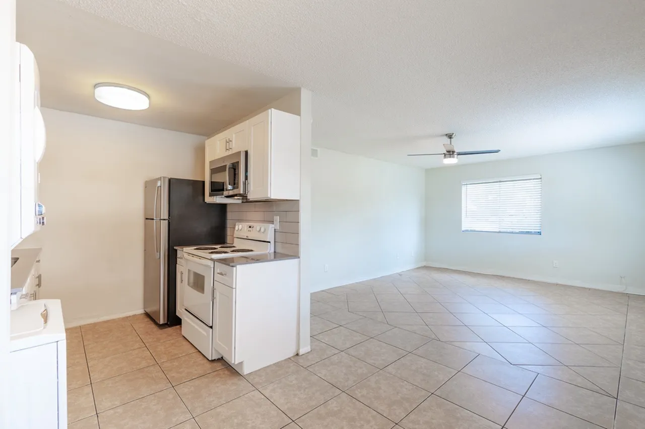 Kitchen with white cabinetry and stainless appliances, wood-style plank flooring, open to spacious living room with ceiling fan and window