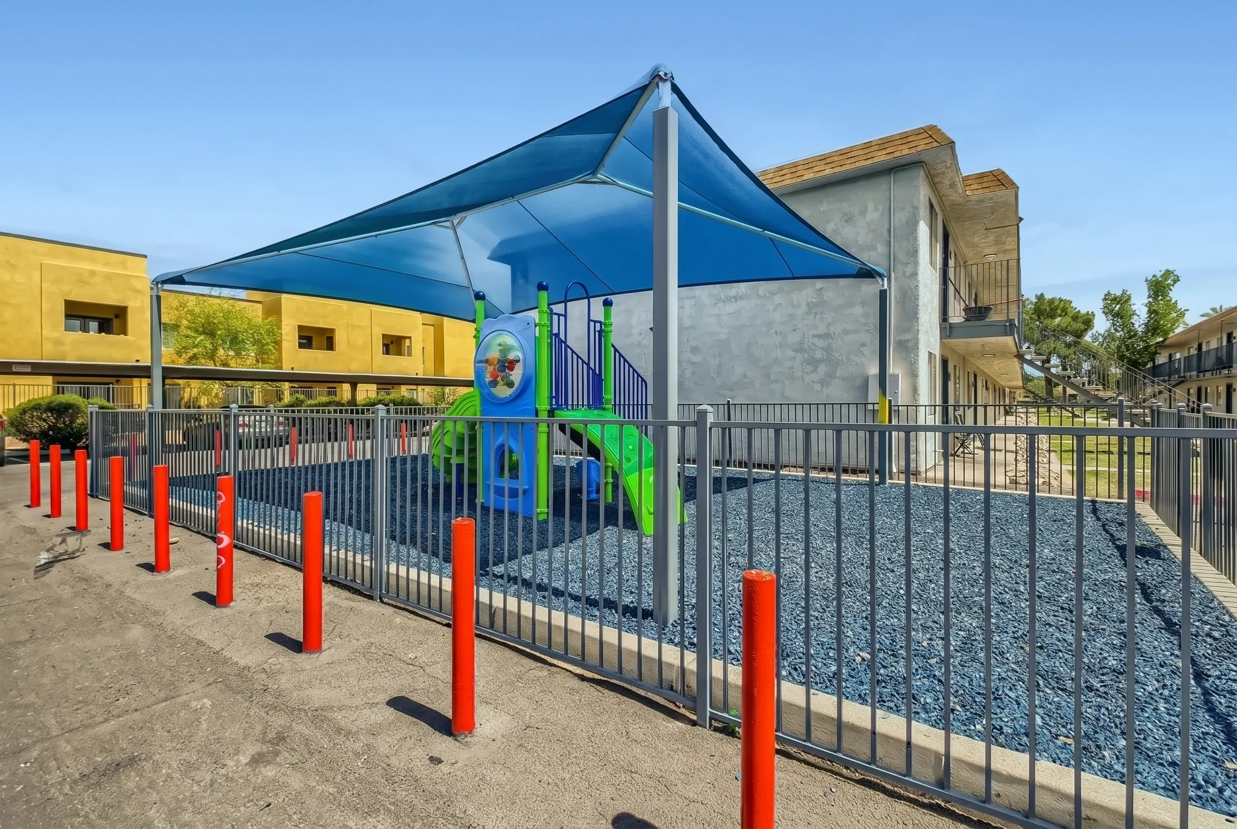 Playground with blue shade canopy, green play structure, red safety bollards, black fencing, gravel surface