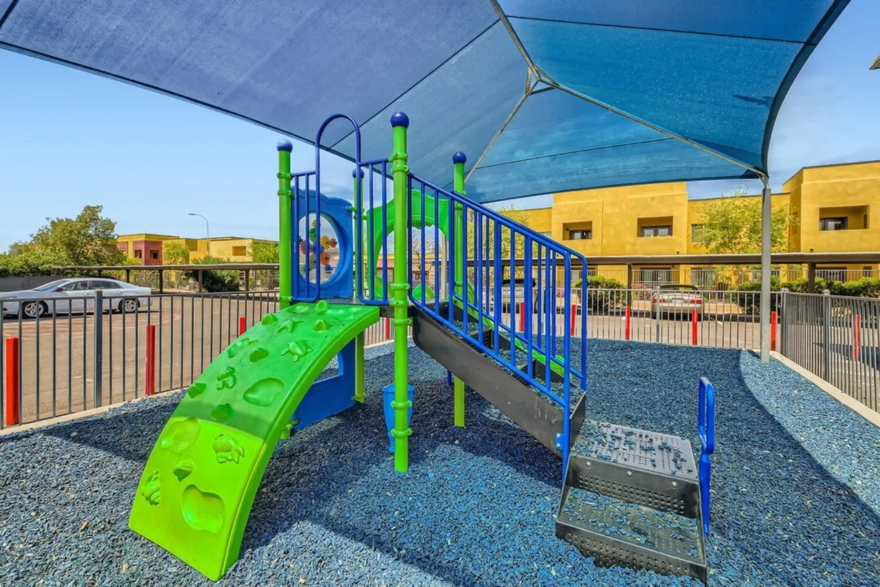 Playground, green slide, blue climbing structure, shade canopy, ground cover, yellow building