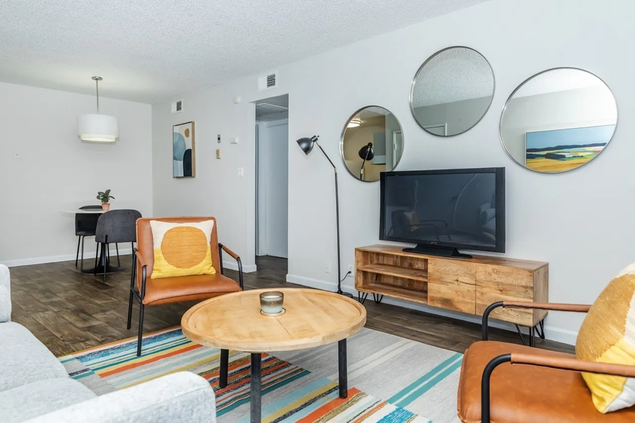 Living room with wood-style plank flooring, orange mid-century chairs, round wood coffee table, wooden TV stand, circular mirrors, black floor lamp