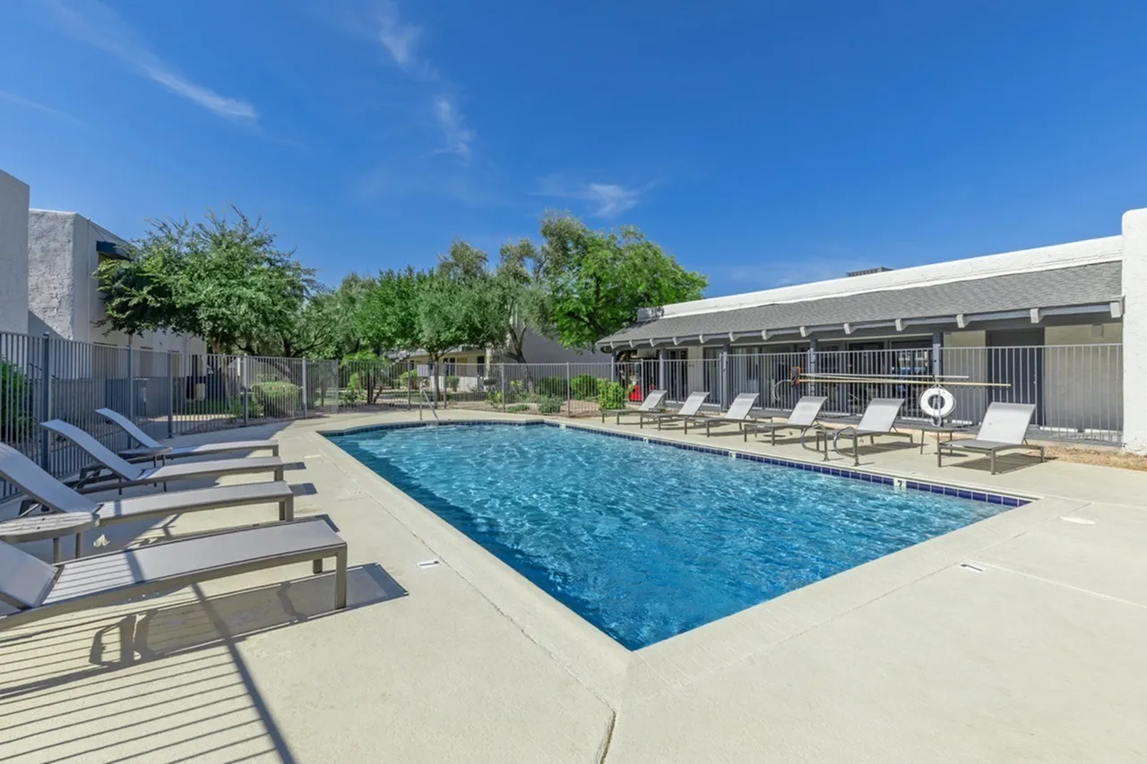 Swimming pool, concrete deck, lounge chairs, metal fencing, building with covered seating area, trees