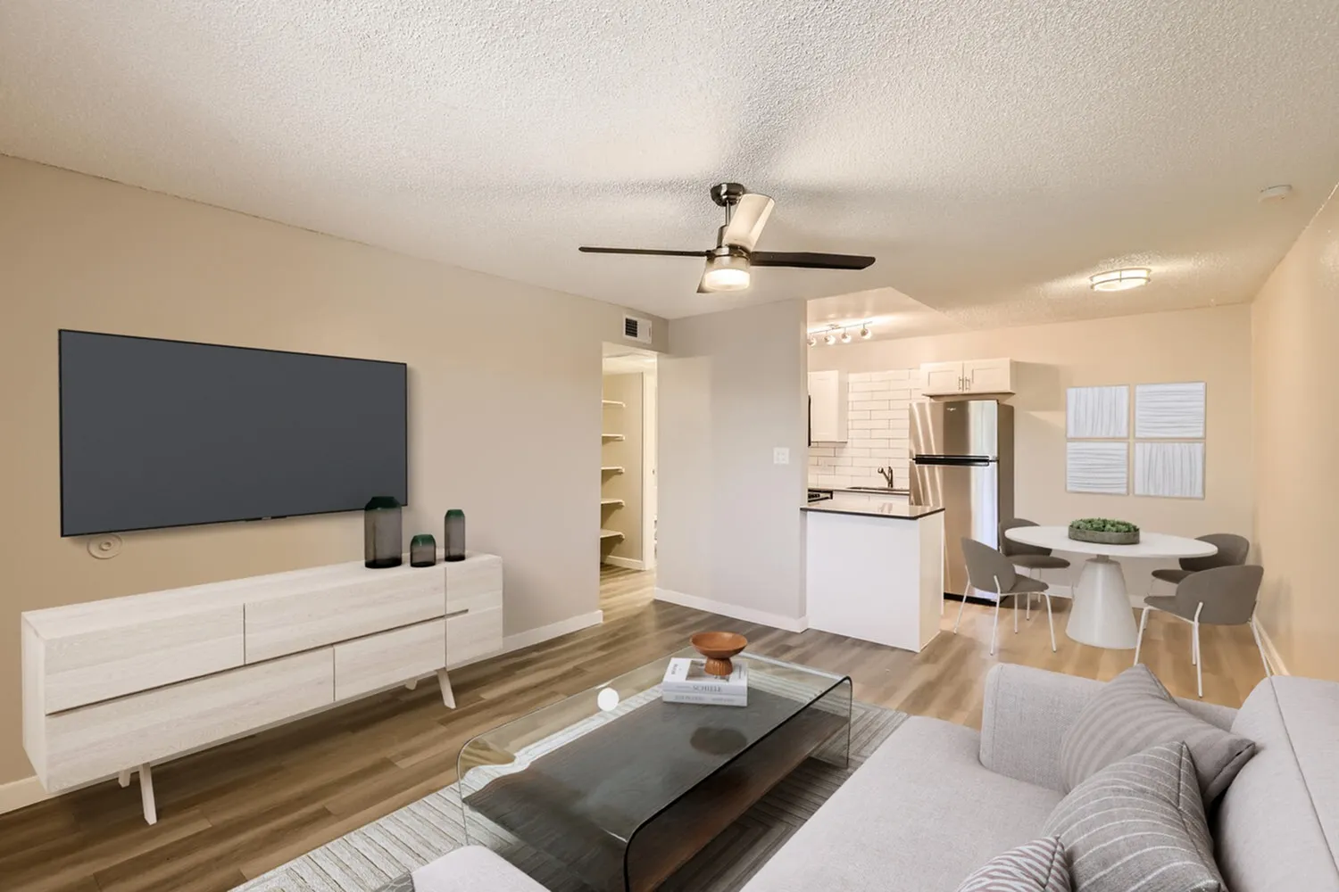 Living room with wood-style plank flooring, ceiling fan, flat-screen TV, white media console, dining area with round table, open kitchen with white cabinetry and stainless appliances