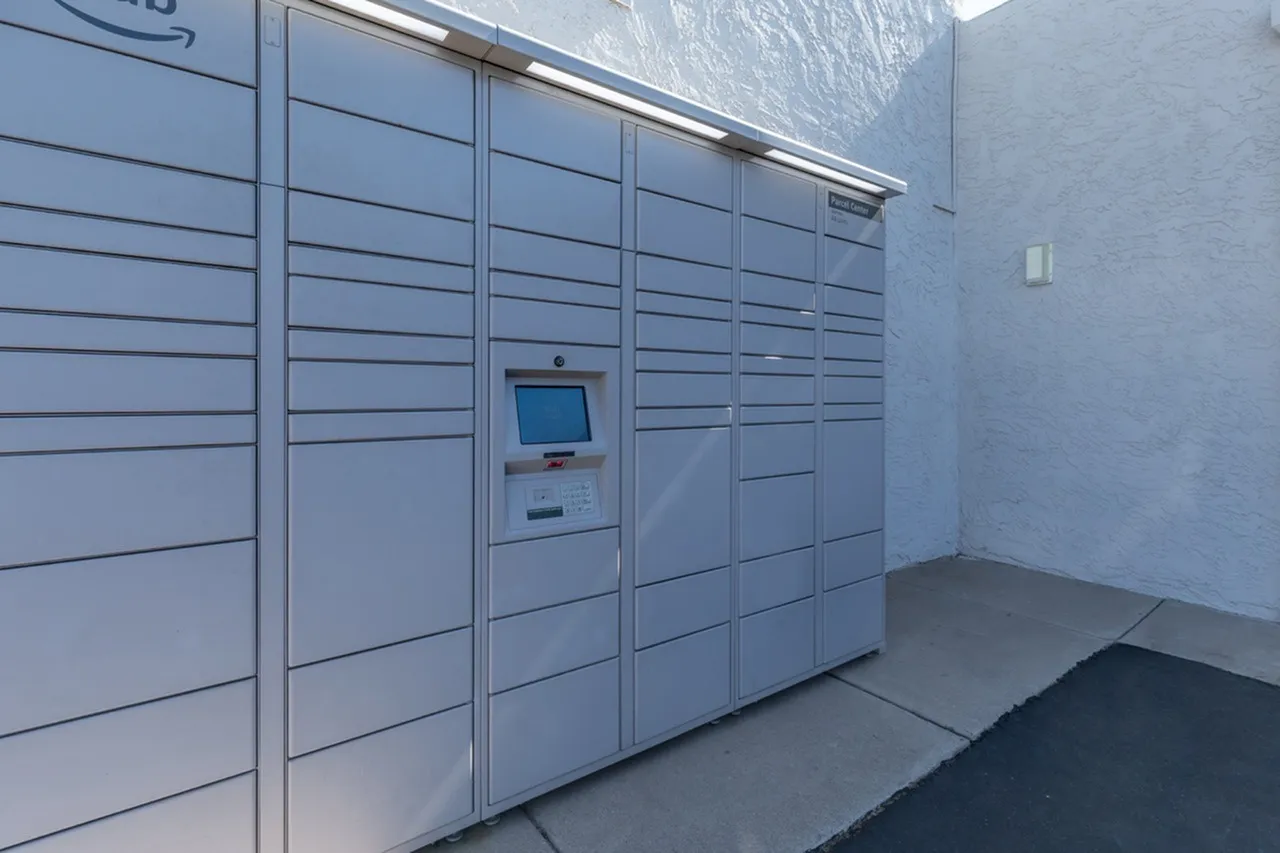 Package locker wall, white metal doors with horizontal lines, digital touchscreen kiosk, concrete flooring