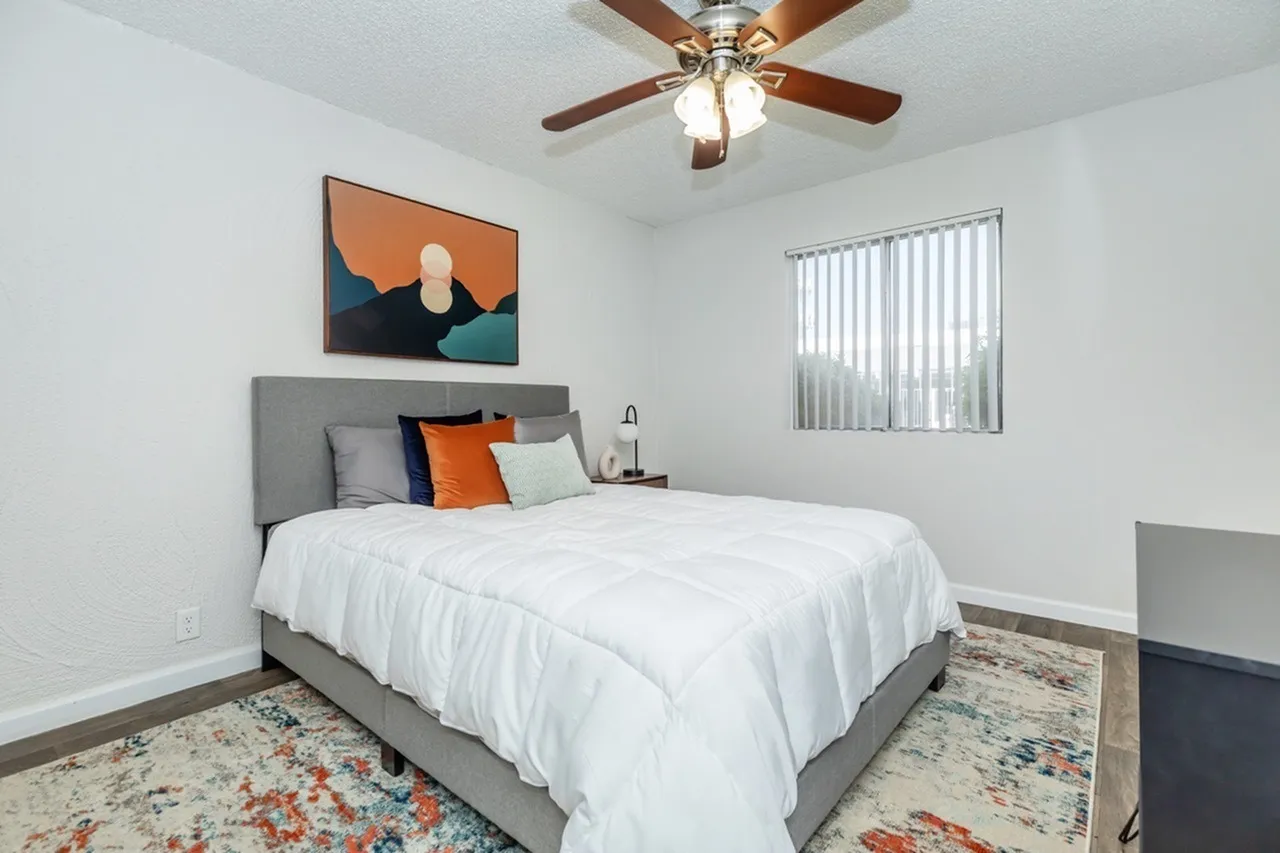 Primary bedroom, gray upholstered headboard, white bedding, orange accent pillow, ceiling fan, wood-style plank flooring