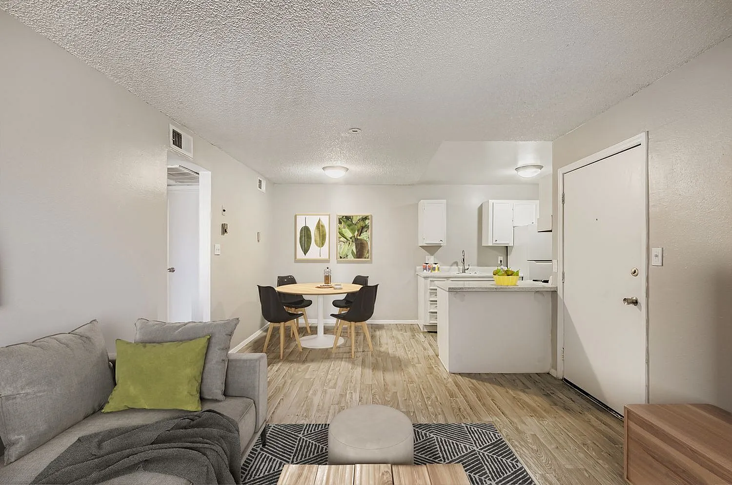 Living room with gray sofa, wood-style plank flooring, dining area, kitchen with white cabinetry, bathroom entry