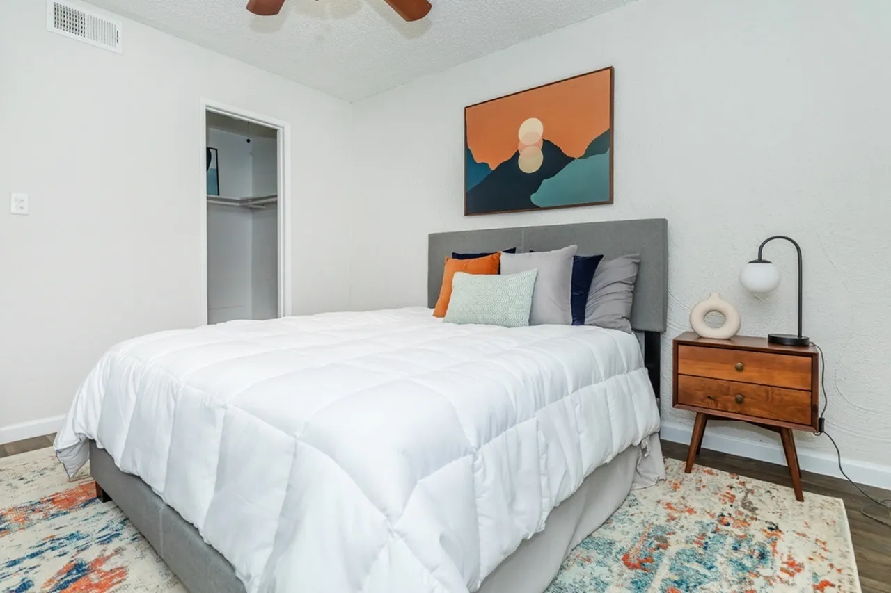 Primary bedroom, gray upholstered headboard, white comforter, wood-style nightstand, abstract wall art, wood-style plank flooring