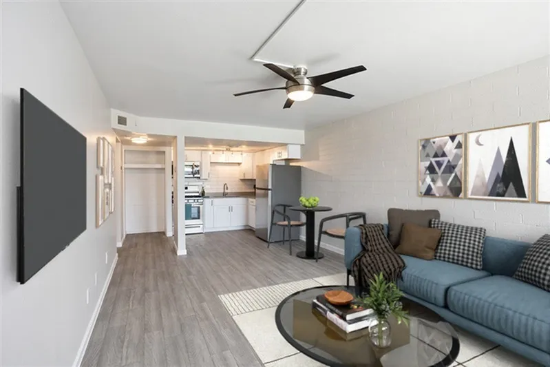Living room and kitchen, wood-style plank flooring, ceiling fan, blue upholstered sofa, round glass coffee table, white cabinetry, stainless appliances, geometric wall art