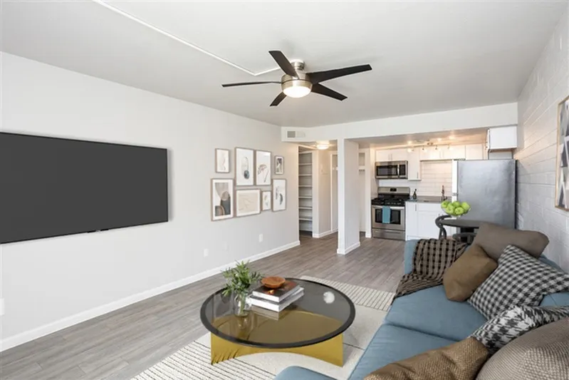 Living room with wood-style plank flooring, ceiling fan, sectional sofa, round coffee table, mounted television, open kitchen with stainless appliances