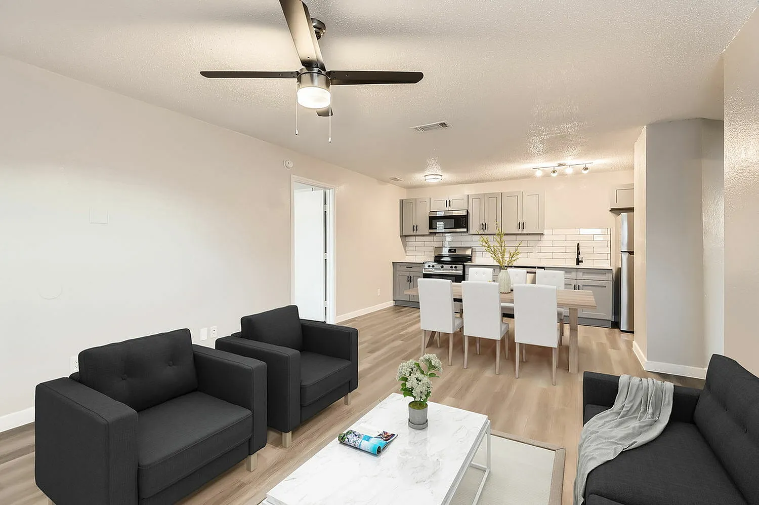Living room with wood-style plank flooring, black upholstered seating, ceiling fan, open to kitchen with white shaker cabinetry and stainless appliances