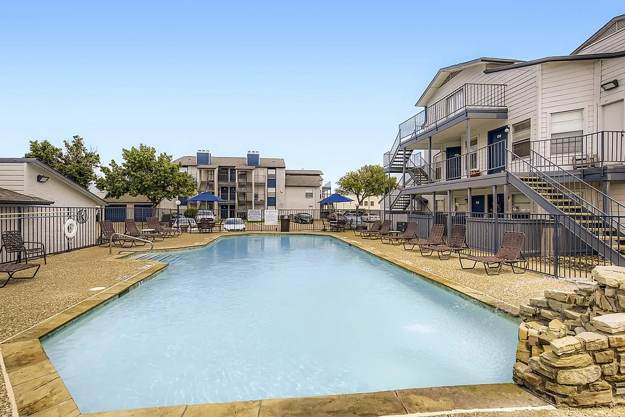 Swimming pool with light blue water, concrete deck, metal railings, lounge chairs, multi-story residential buildings