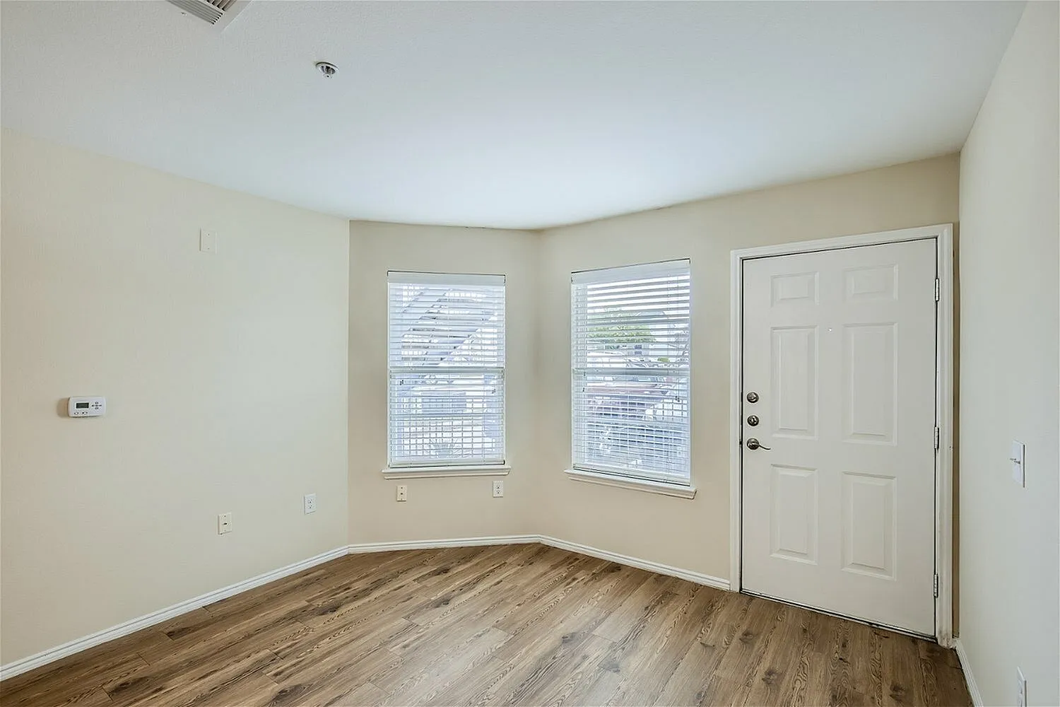 Living room, wood-style plank flooring, windows with blinds, white entry door, cream walls