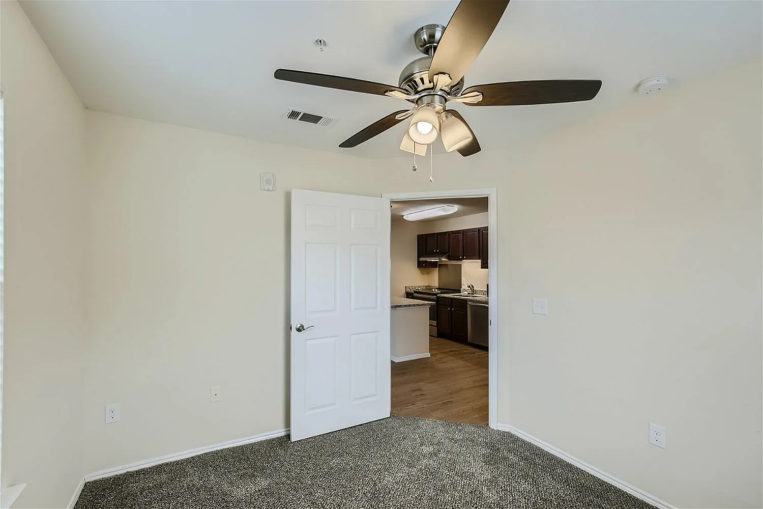 Bedroom, gray carpet flooring, ceiling fan with lights, open doorway to kitchen