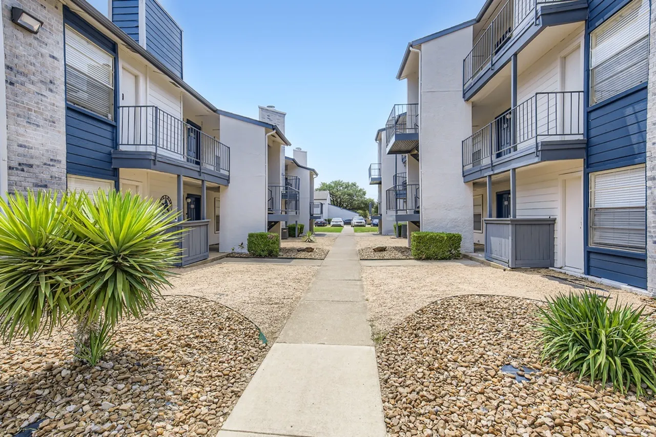 Courtyard pathway, multi-story residential buildings, blue metal railings, mature landscaping, gravel grounds