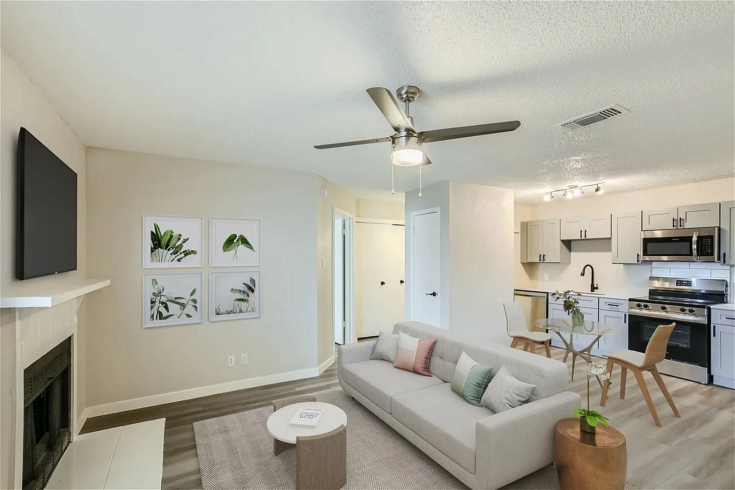 Living room with wood-style plank flooring, ceiling fan, fireplace, wall-mounted TV, kitchen with stainless appliances