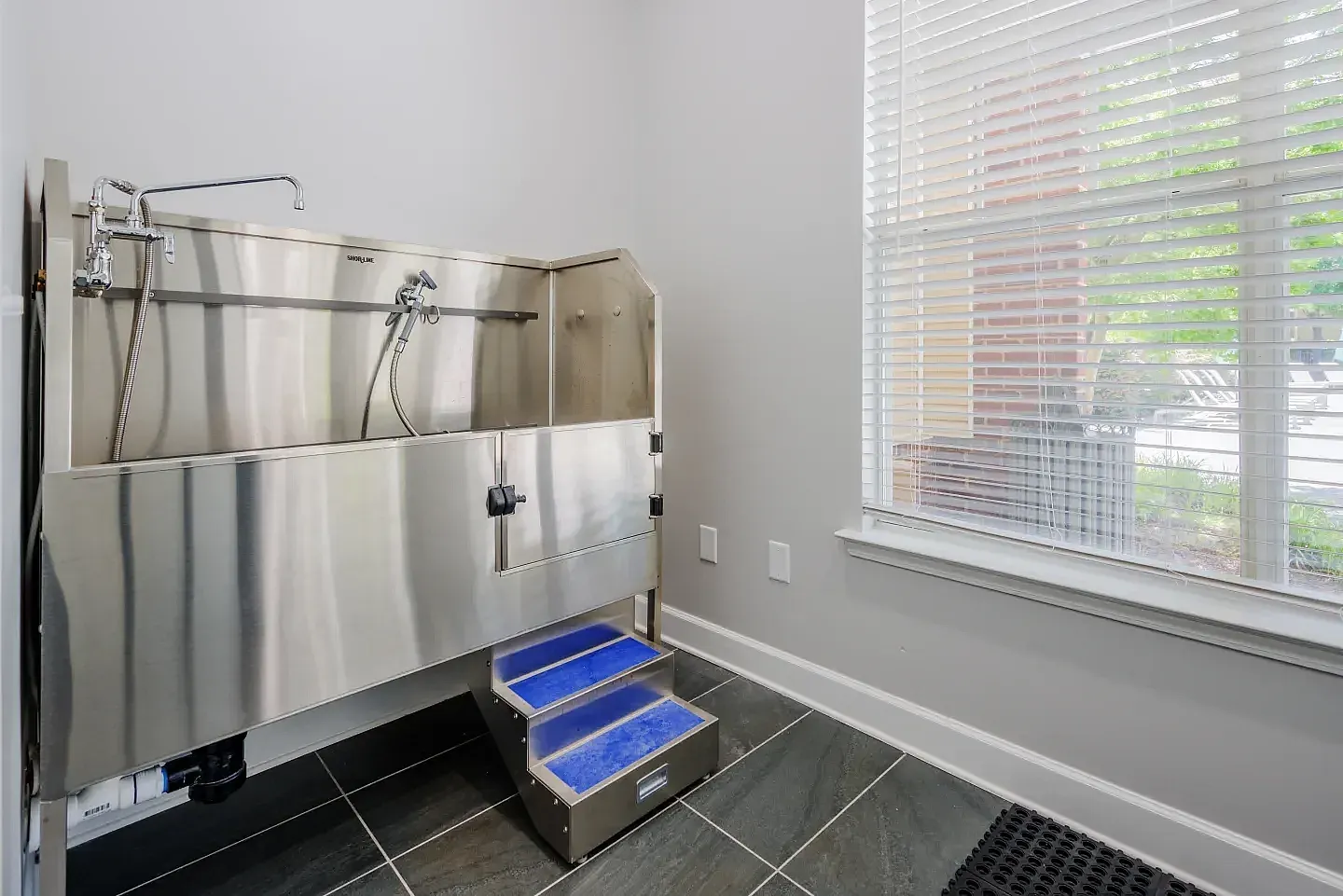 Dog wash room, stainless steel elevated tub, blue step stool, dark tile flooring, horizontal blinds
