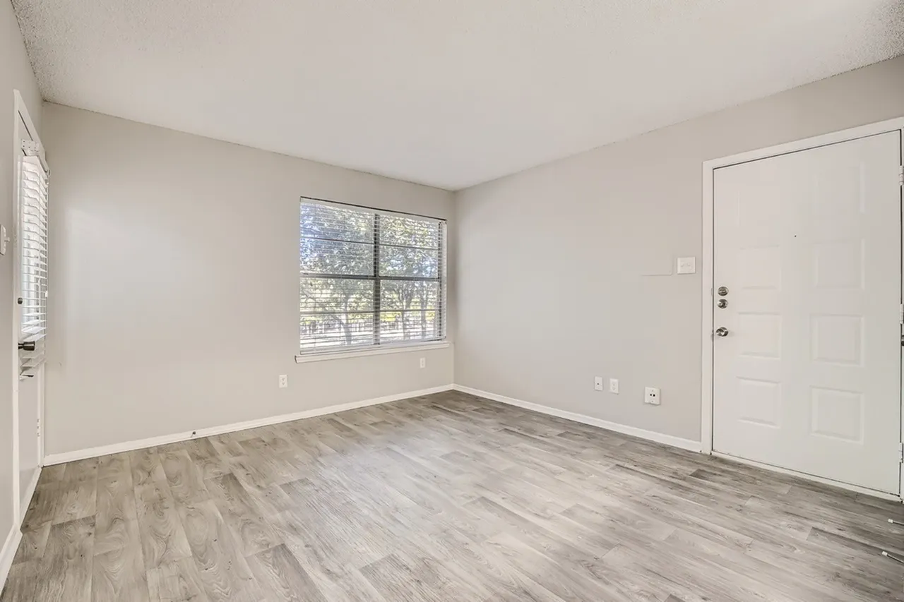 Apartment room, wood-style plank flooring, large window, white walls, entry door