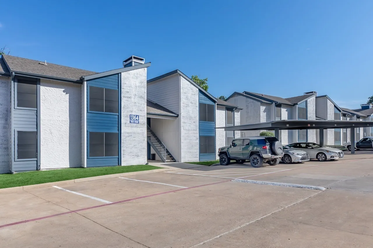 Modern apartment complex with white and blue facade, multiple buildings, parked vehicles, clear sky, landscaping