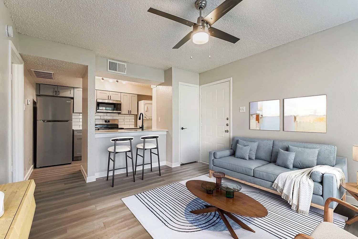 Open concept living room and kitchen, wood-style plank flooring, gray upholstered sofa, white cabinetry, stainless appliances