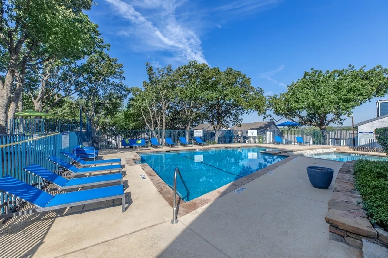 Resort-style swimming pool, lounge chairs, blue umbrellas, mature trees, clear blue sky