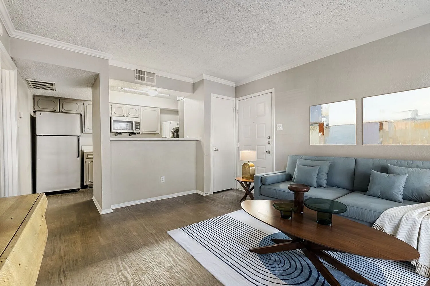 Living room with blue sofa, wood-style plank flooring, striped area rug, open kitchen with white cabinetry