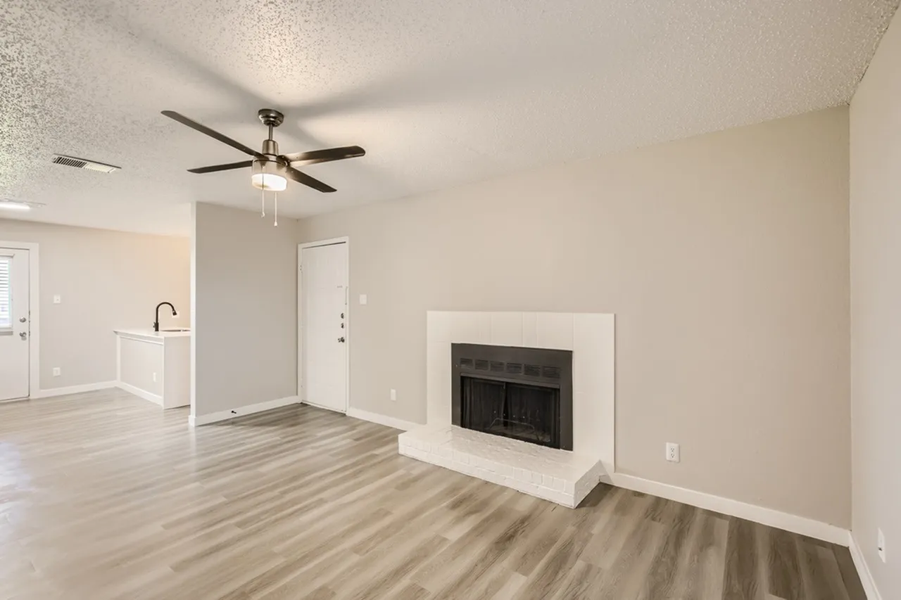 Living room, wood-style plank flooring, ceiling fan, fireplace with white surround, kitchen visible