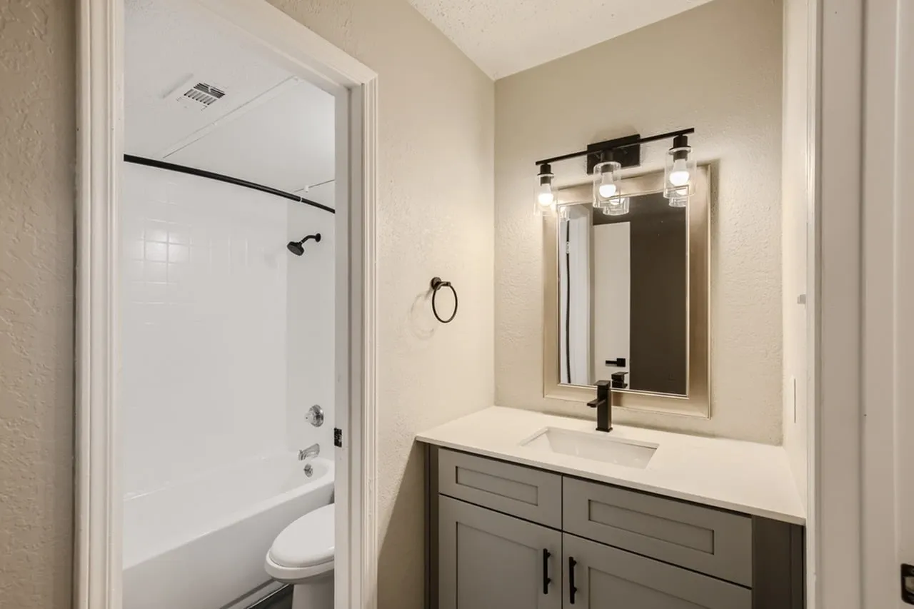 Bathroom, gray vanity cabinetry, white quartz countertop, framed mirror with lighting, glass shower enclosure
