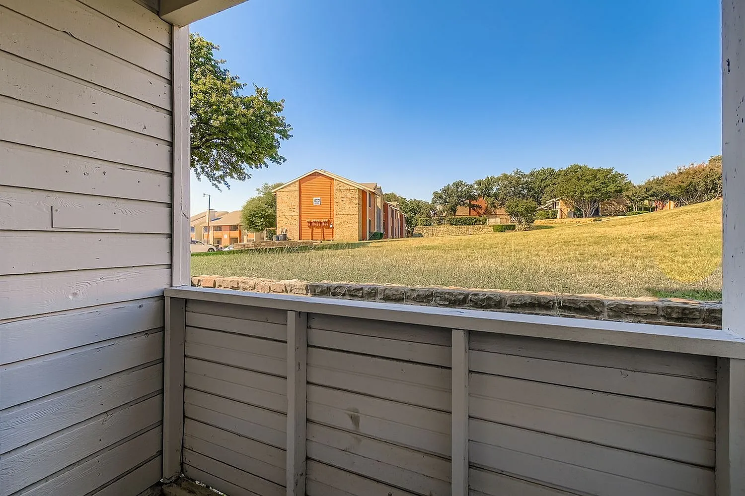 Balcony with horizontal railing, brick building, green lawn, trees, clear sky
