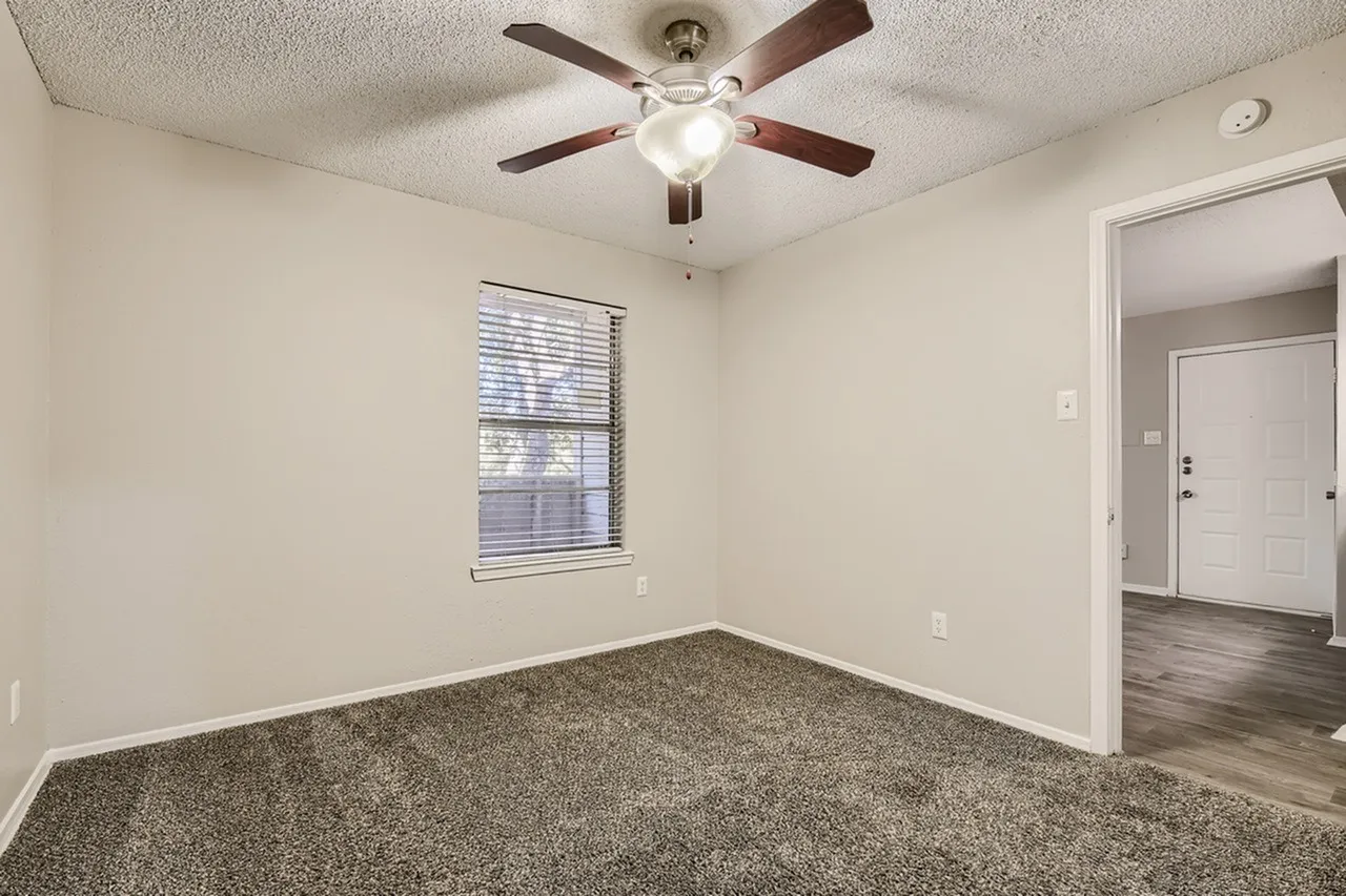 Bedroom, gray carpet flooring, ceiling fan with light, window with blinds, entry door to hallway