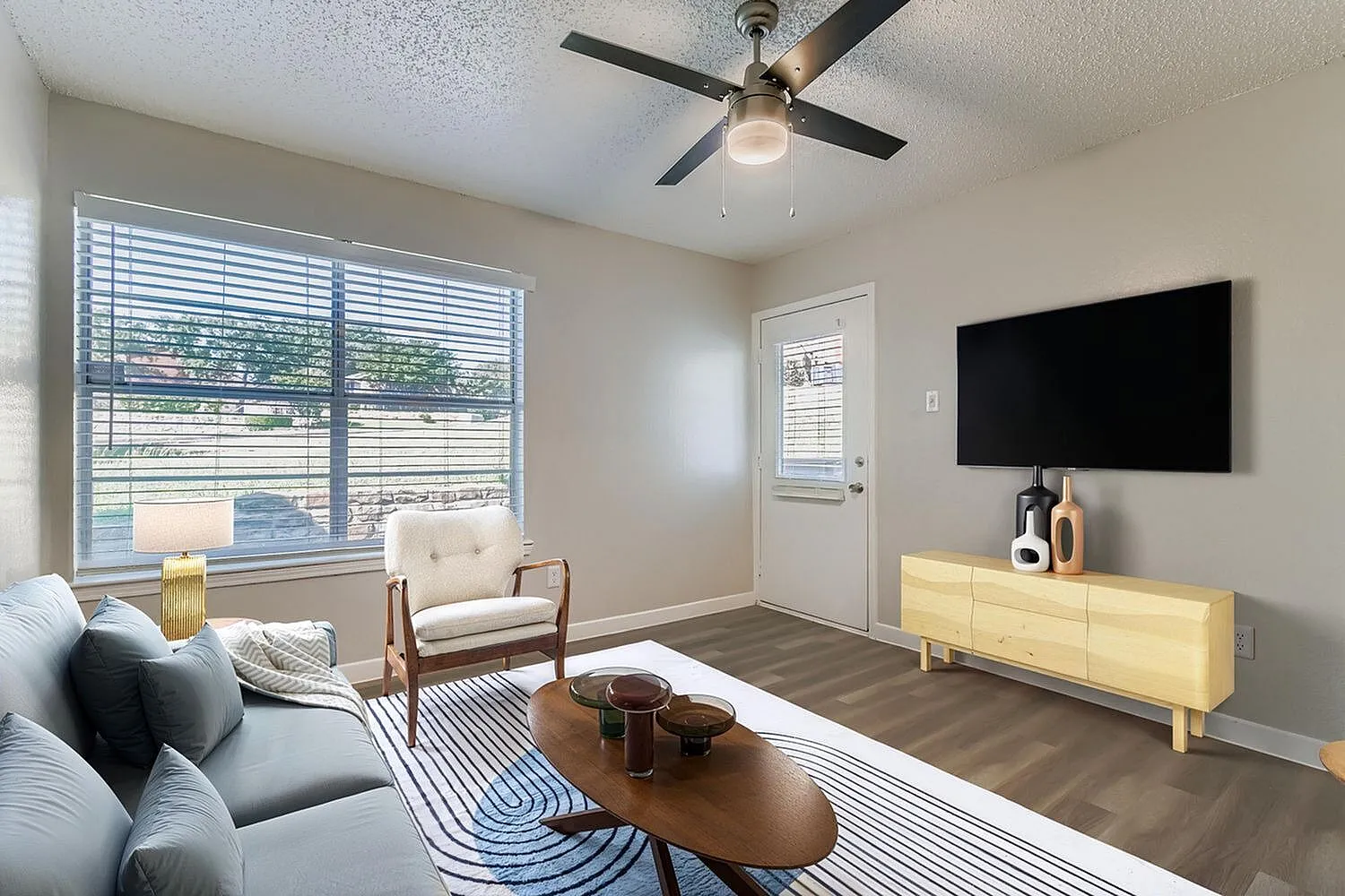 Living room, wood-style plank flooring, ceiling fan, flat-screen TV, entry door