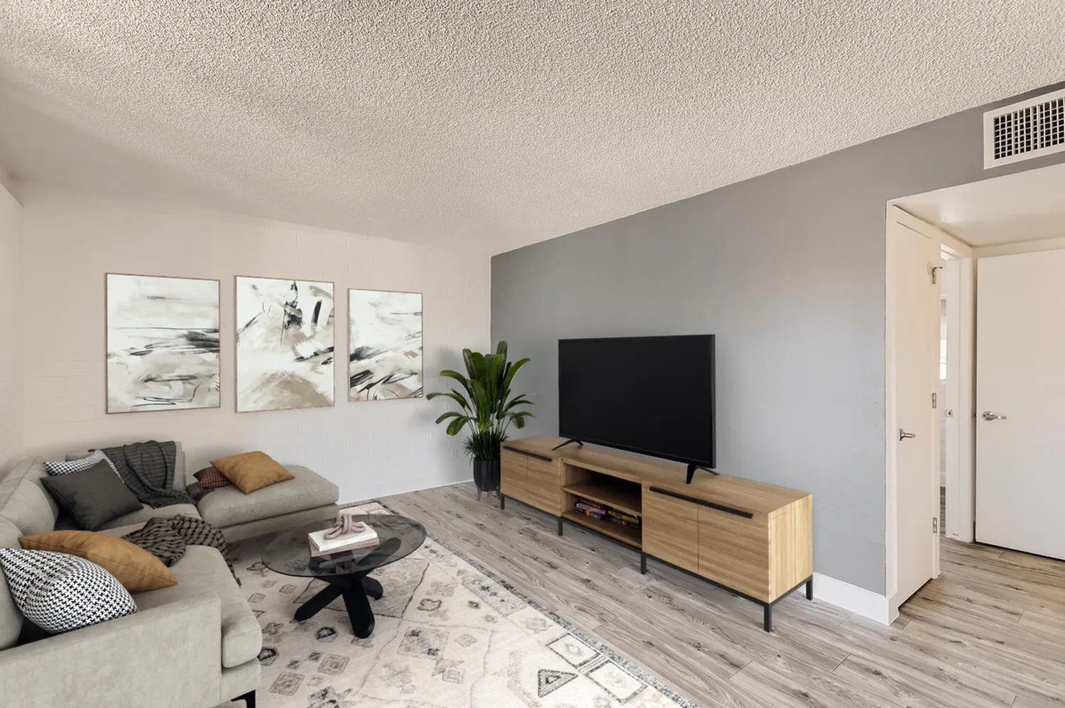 Living room with wood-style plank flooring, gray accent wall, TV on wood media console, gray sofa with throw pillows, white area rug, framed wall art