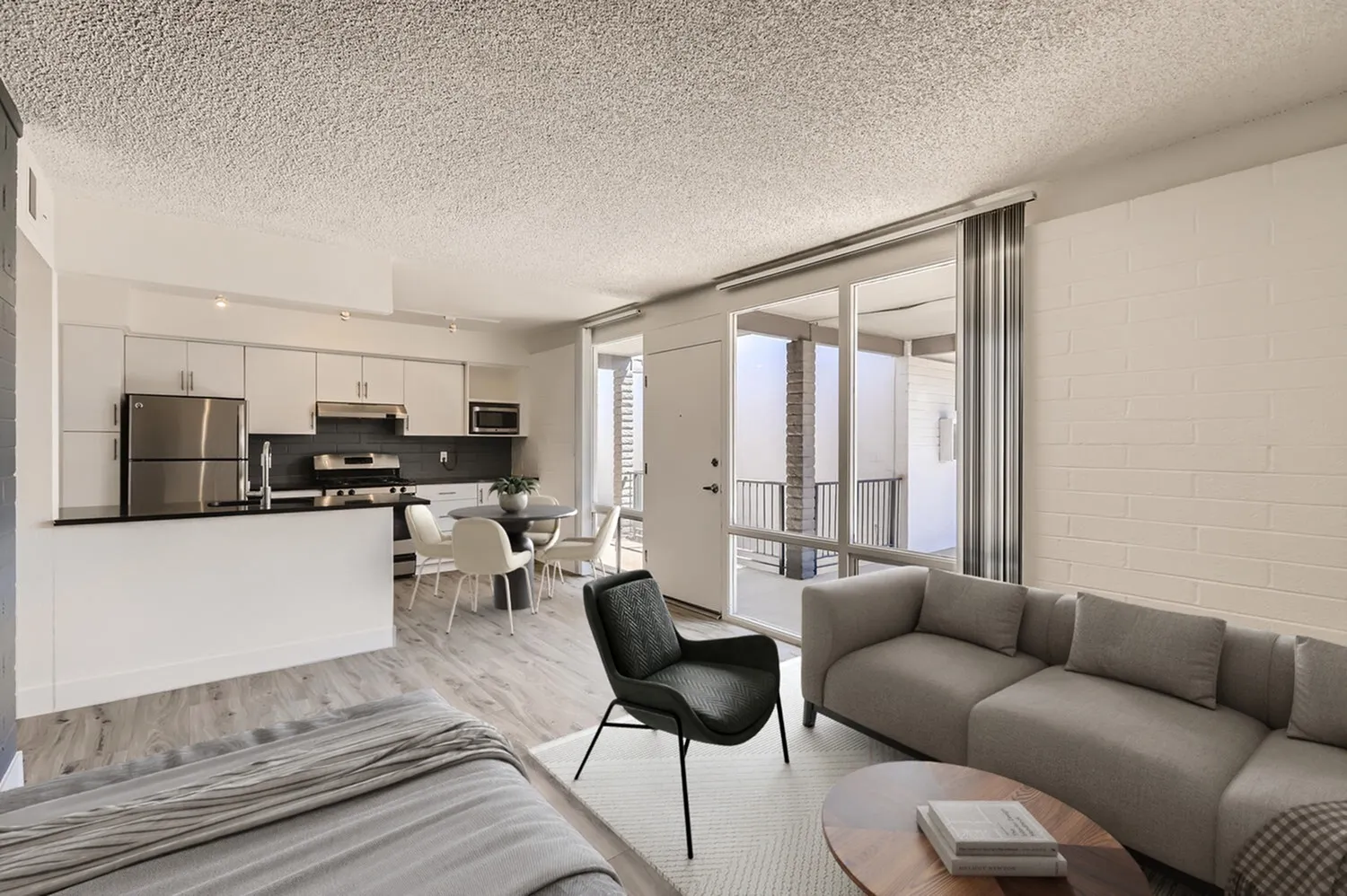 Open-concept studio, wood-style plank flooring, white cabinetry, stainless appliances, gray upholstered sofa, sliding glass patio door