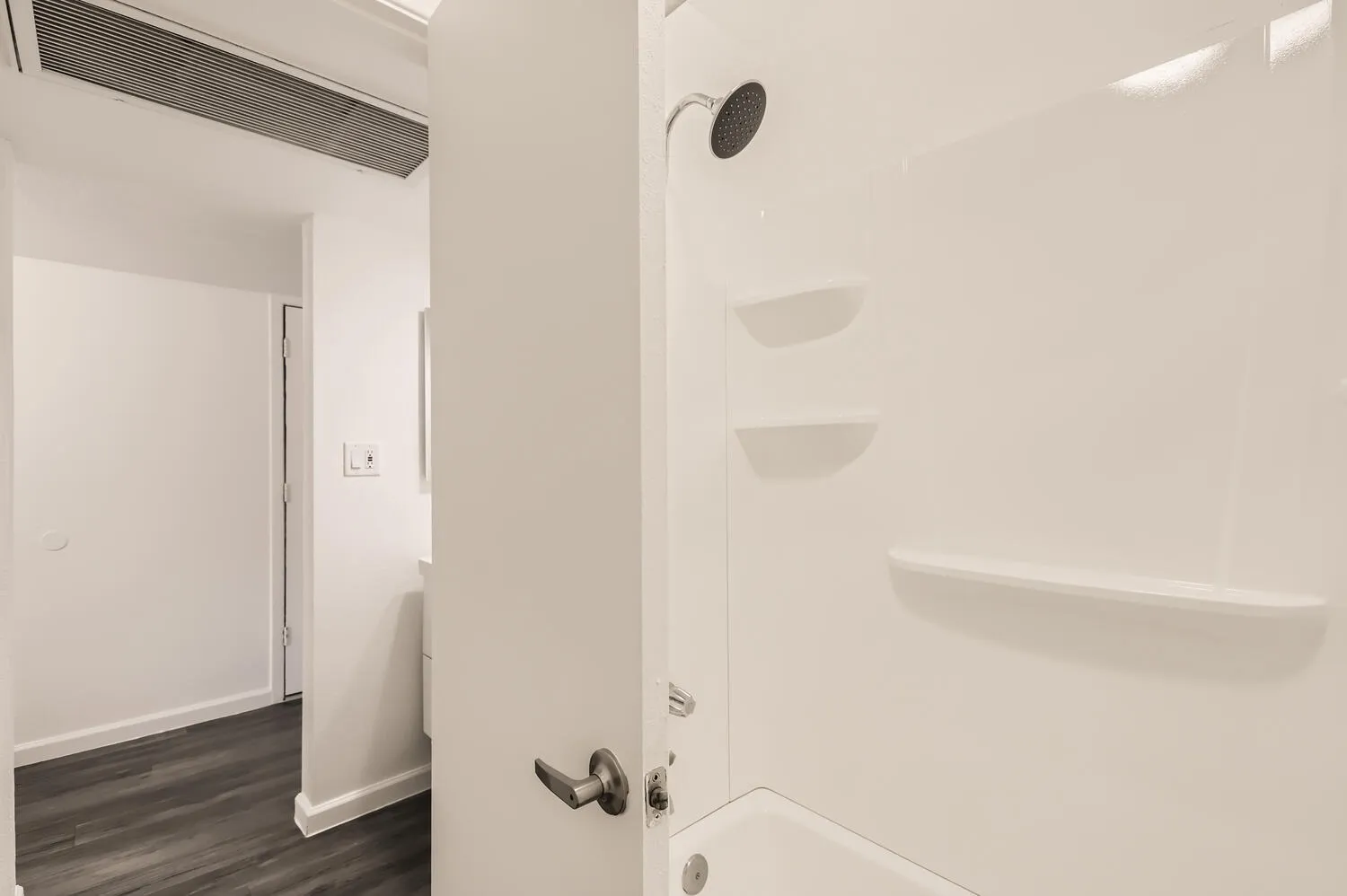 Bathroom, white tiled shower wall, built-in shelving, dark wood-style plank flooring, overhead ventilation