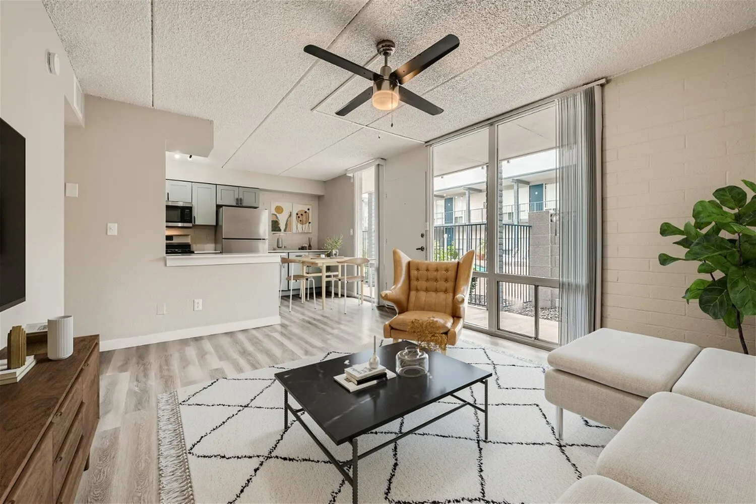 Living room with wood-style plank flooring, ceiling fan, black coffee table, tan upholstered sofa, geometric area rug, open kitchen with white cabinetry, glass sliding doors to balcony