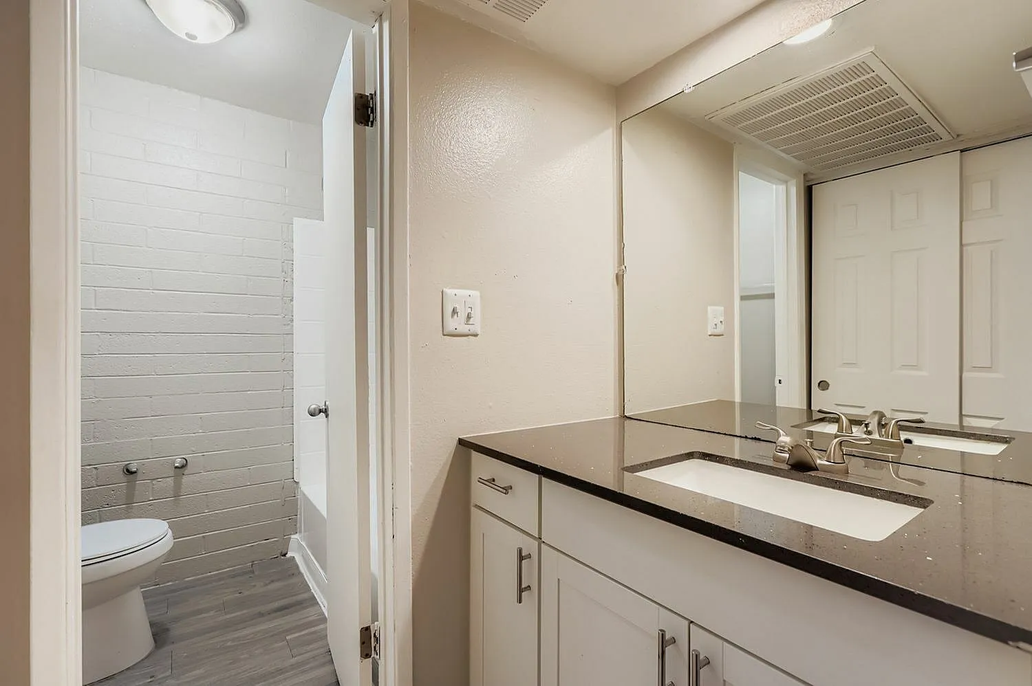 Bathroom, white cabinetry, black countertops, large mirror, glass shower enclosure, wood-style plank flooring