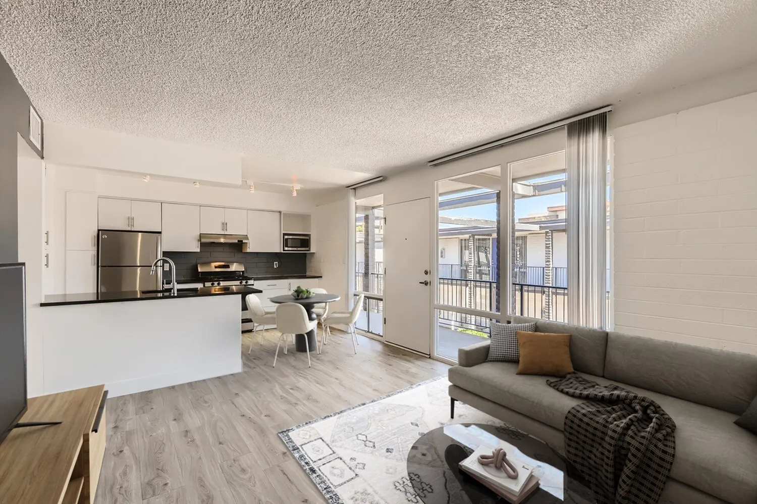 Open concept living room and kitchen, wood-style plank flooring, stainless steel appliances, gray sofa, sliding glass doors to balcony