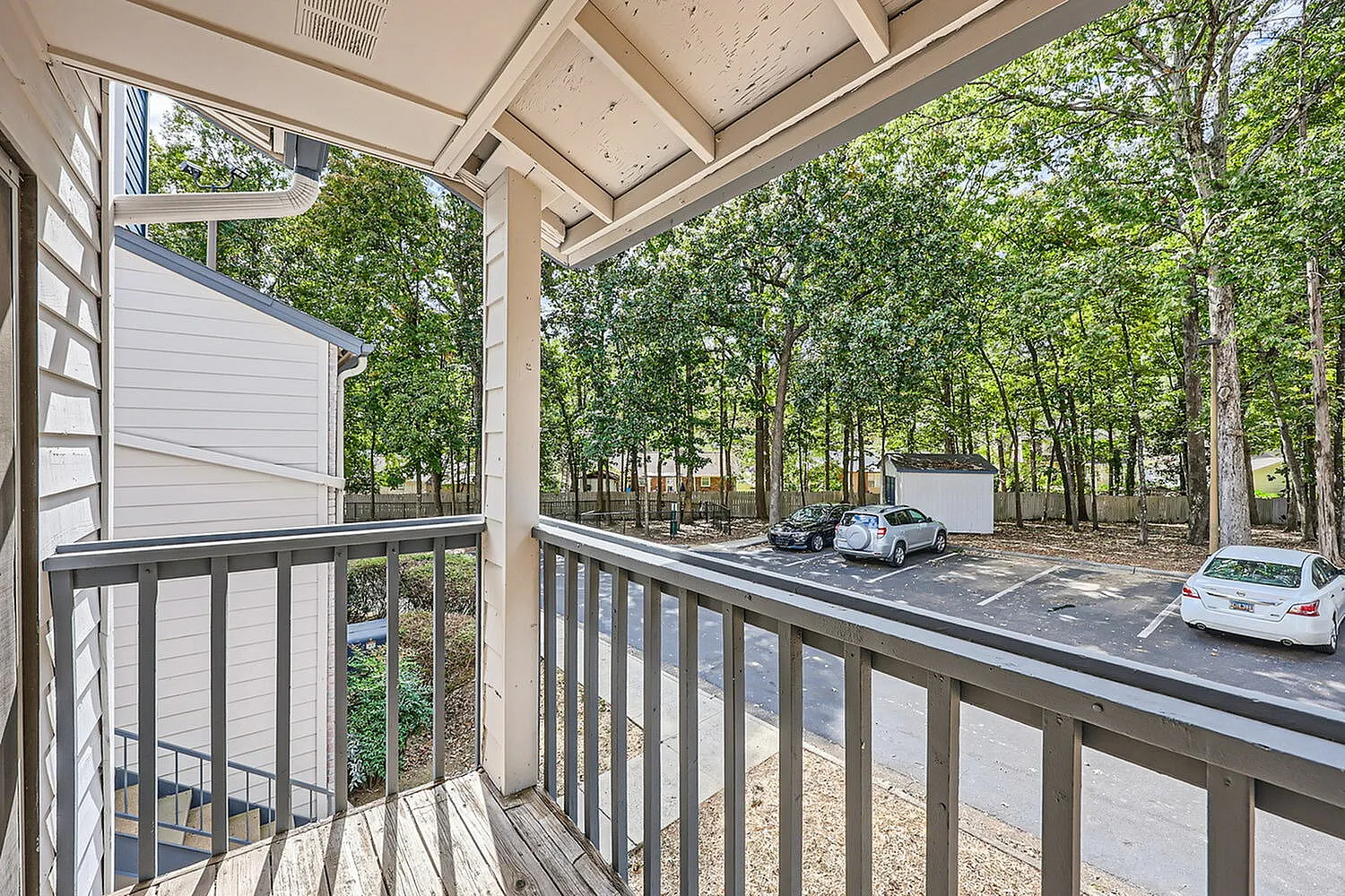 Covered porch with railing, wooded driveway view, parked vehicles